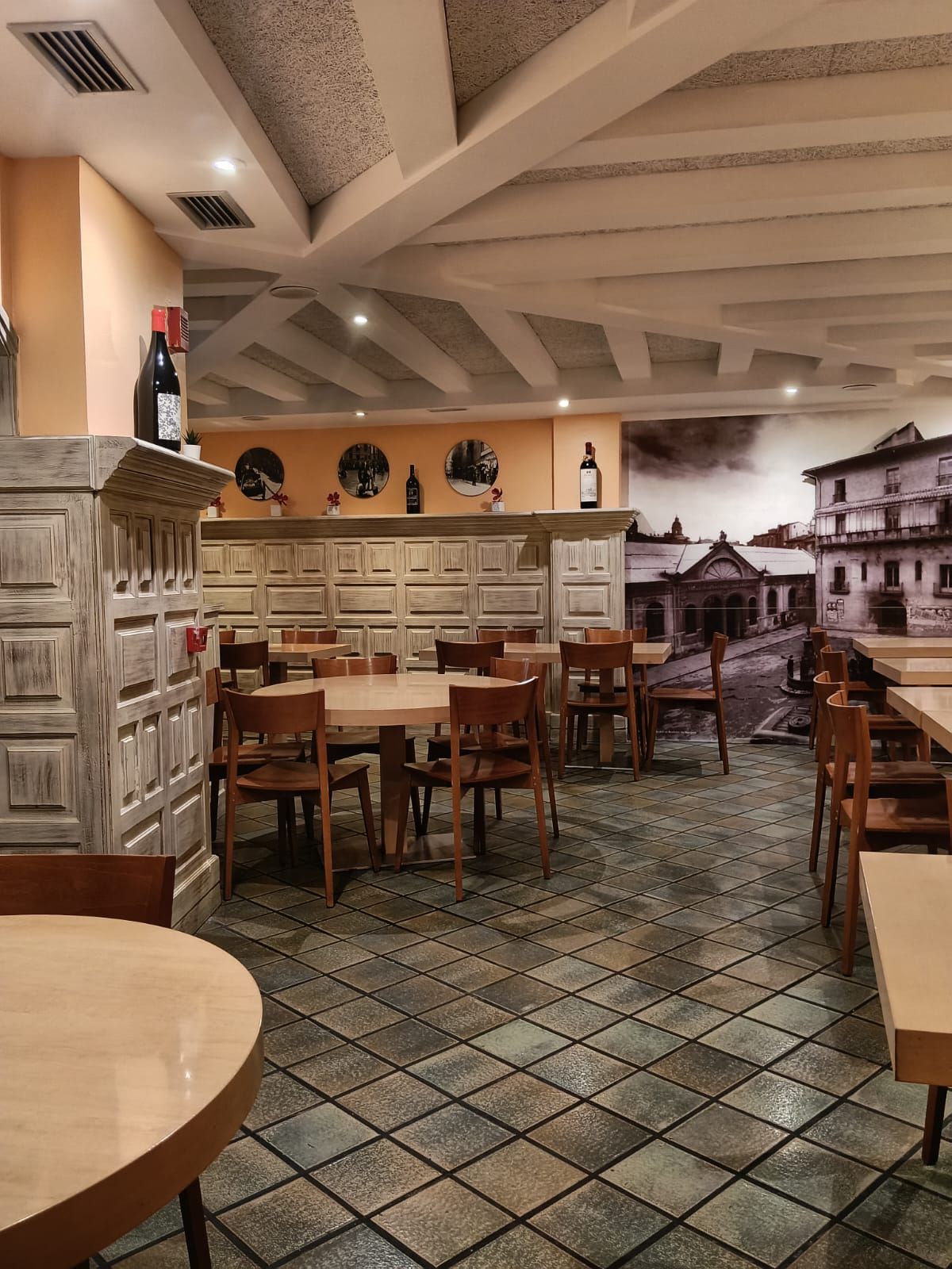El comedor con las mesas de madera clara, la pared de casetones y un gran mural en blanco y negro de una calle antigua de Pamplona.