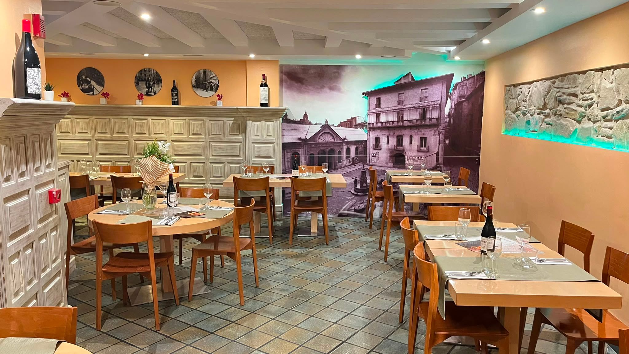 El comedor con las mesas puestas, el mural en blanco y negro del casco viejo y la tira de luz turquesa sobre el muro de piedra.