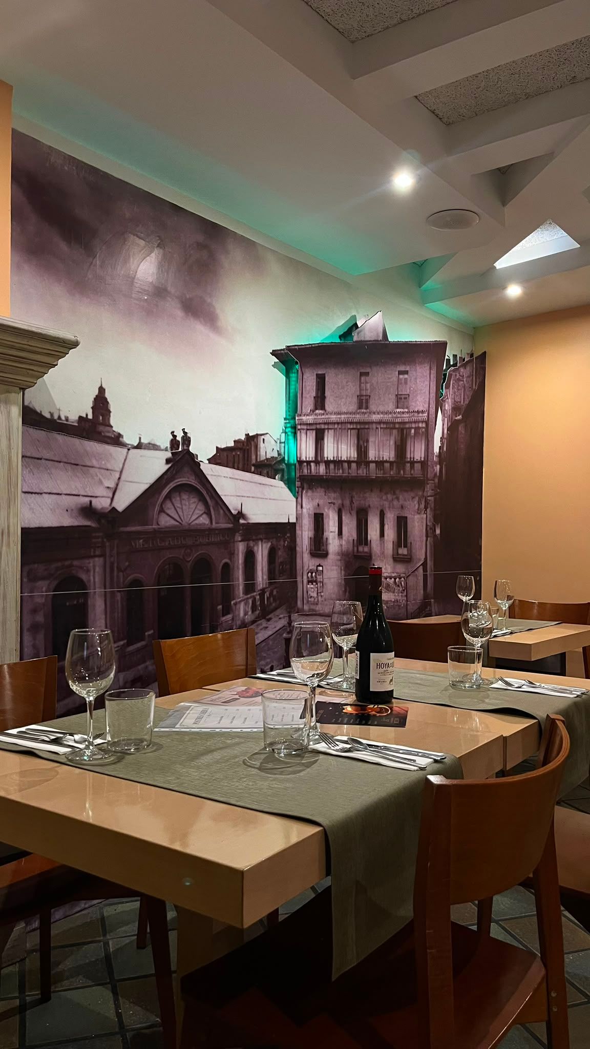Una mesa para dos junto al mural, con copas, cubiertos y una botella de vino tinto.