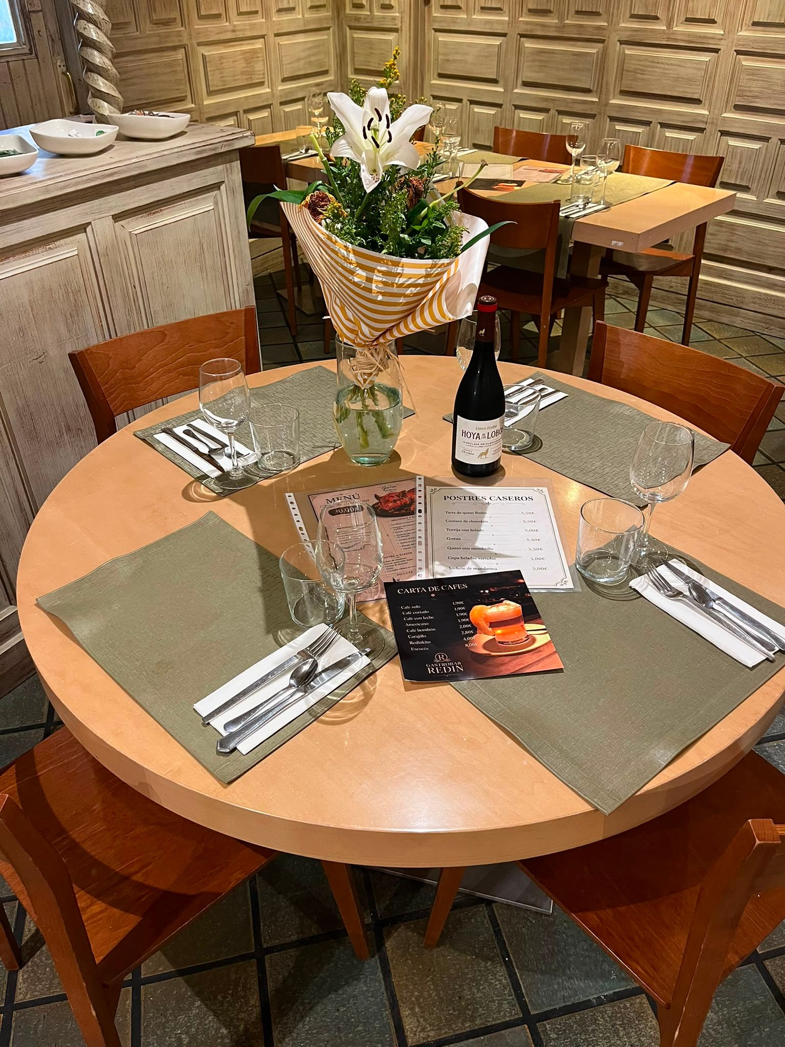 La mesa redonda puesta para cuatro, con flores, una botella de vino y las cartas de postres y de cafés.