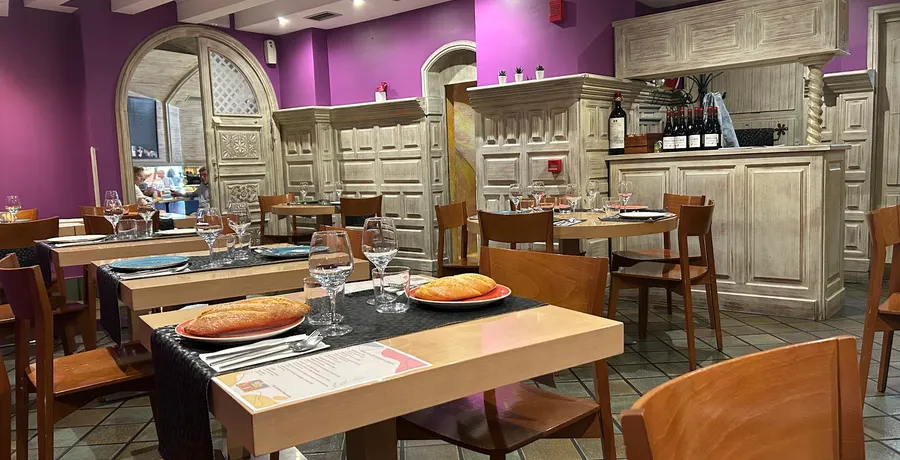 El comedor montado para el servicio, con panes en platos rojos, copas y cubiertos; al fondo, la pared morada, el mueble de madera clara y varias botellas de vino sobre la barra.