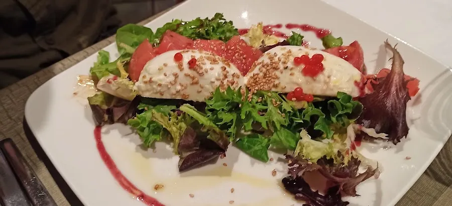 Un plato cuadrado blanco con ensalada de brotes, tomate y dos quenelles de queso cremoso con sésamo y perlas rojas, sobre un trazo de salsa de frutos rojos.