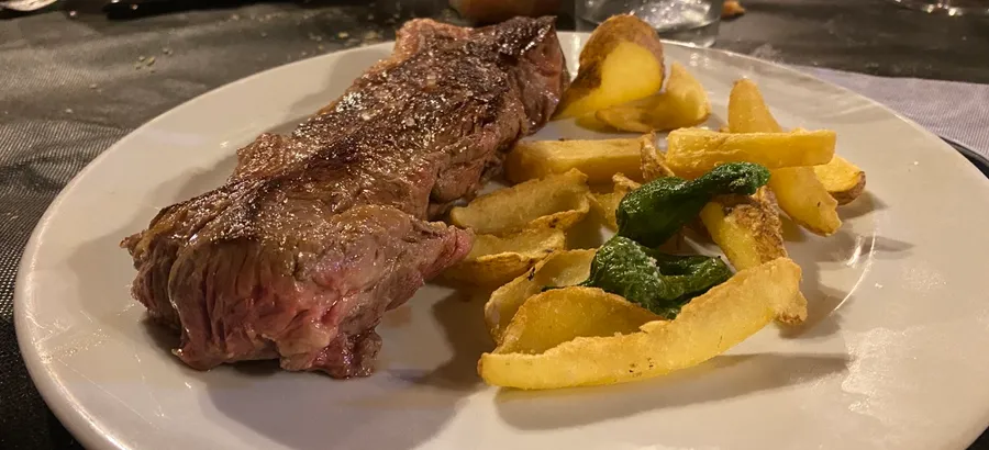 Un plato blanco con un corte de carne a la parrilla, patatas fritas gruesas y un pimiento verde frito.
