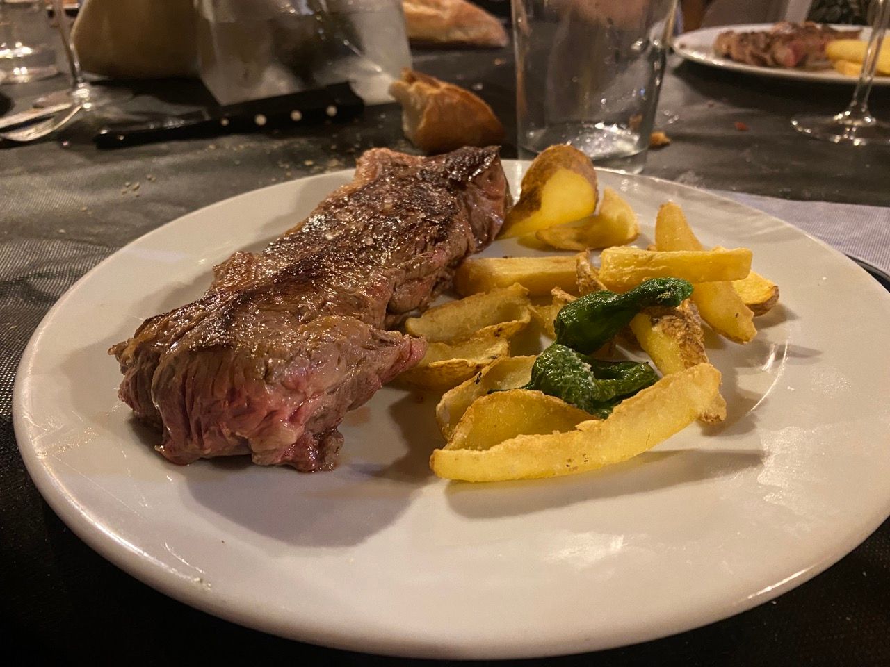 Un plato blanco con un corte de carne a la parrilla, patatas fritas gruesas y un pimiento verde frito.