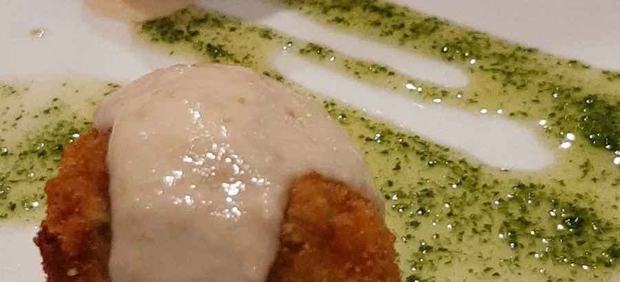 Dos bolas rebozadas y fritas cubiertas de una crema clara, sobre un plato blanco con un hilo de aceite verde de hierbas.