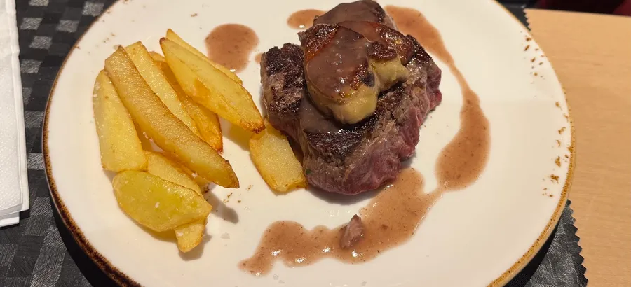 Un medallón de carne con foie y salsa oscura, acompañado de patatas fritas gruesas, en un plato de loza clara.