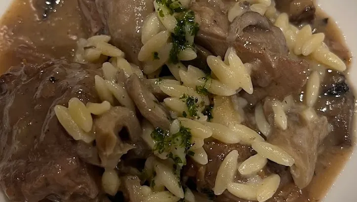 Un plato hondo blanco con carne guisada en su salsa, setas y pasta menuda, espolvoreado con perejil.