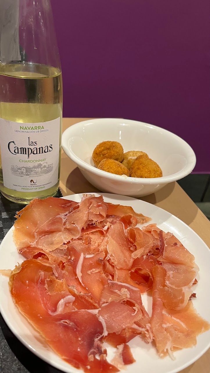 Una botella de vino blanco de Navarra junto a un cuenco de croquetas y un plato de jamón cortado a cuchillo, sobre la pared morada del comedor.