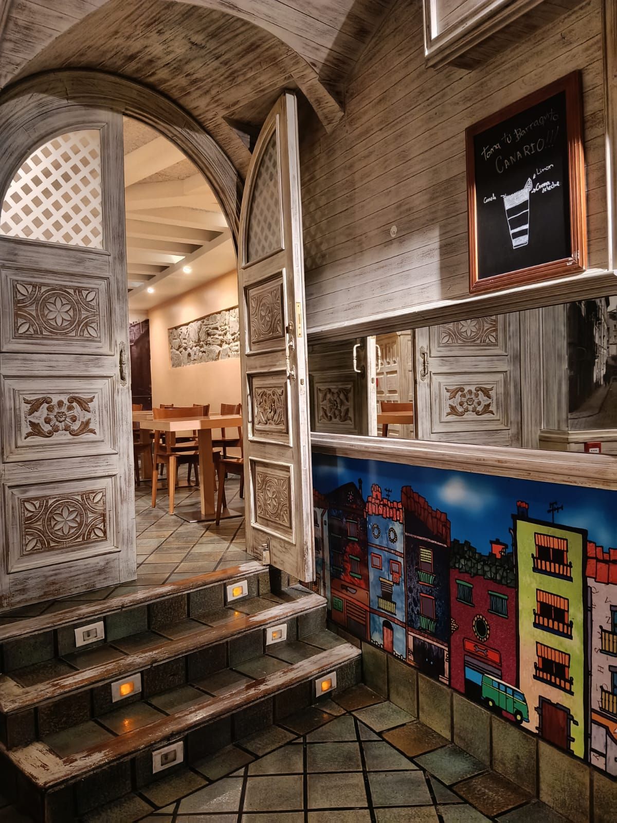 La entrada por dentro: el arco de piedra, la puerta tallada abierta, los escalones iluminados y el mural de casas de colores que sube hacia el comedor.