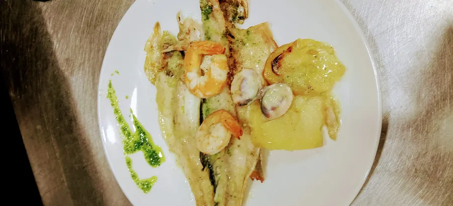 Lubina al horno abierta entera, sobre patatas panaderas y con gambones y almejas por encima, con un hilo de aceite verde de hierbas.