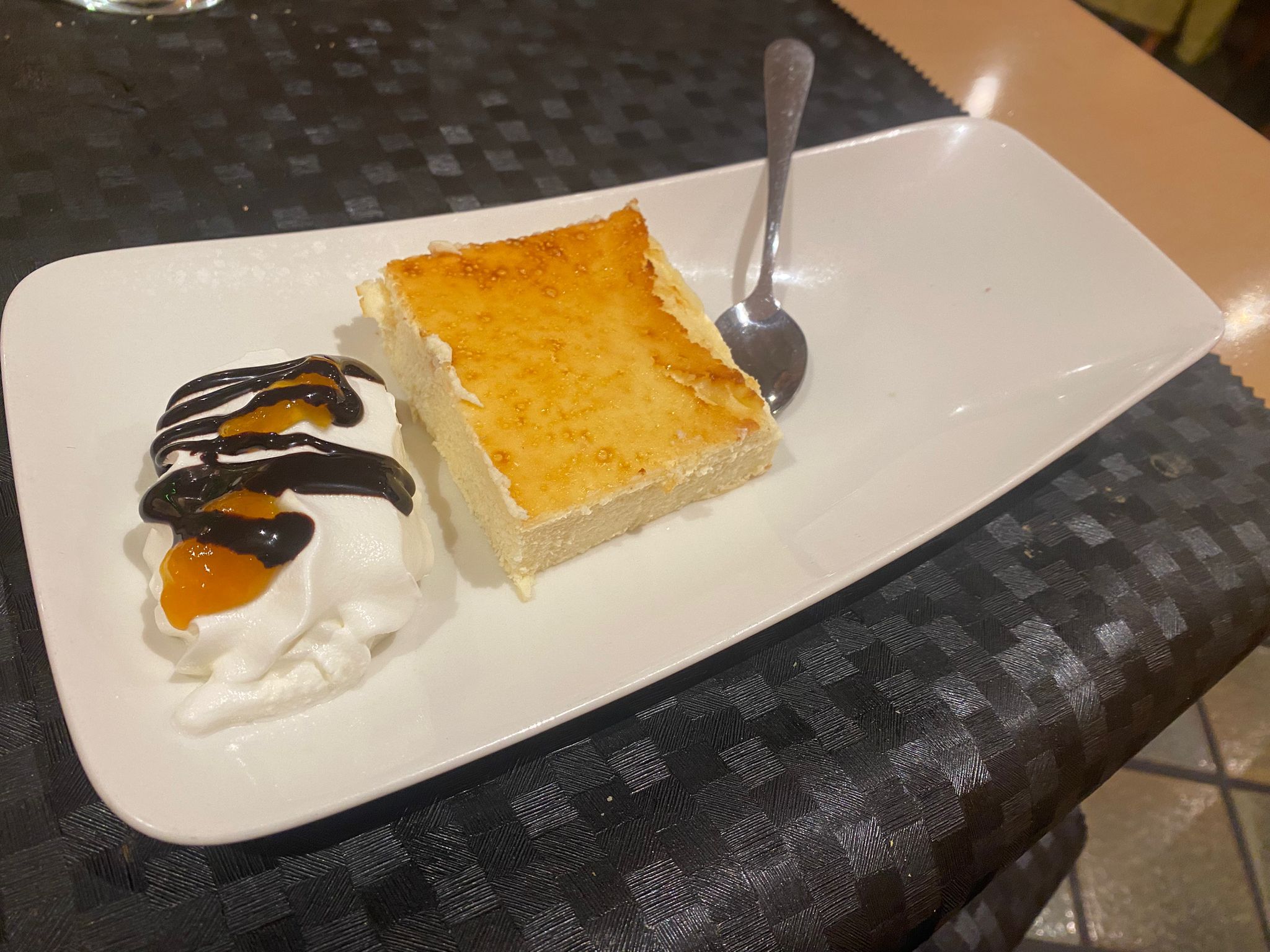 Una porción de tarta de queso con la superficie dorada, servida en un plato alargado con helado y sirope de chocolate.
